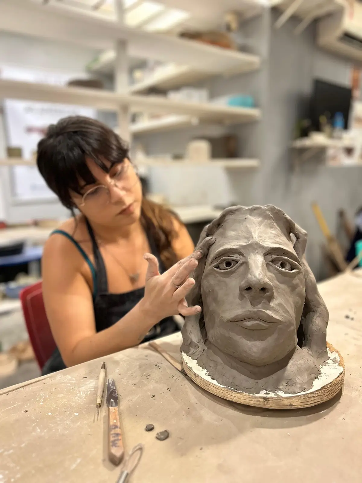 Aluna modela o rosto de um busto de argila na bancada do ateliê.